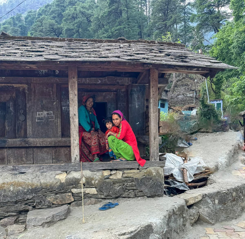 שתי נשים וילד יושבים בפתח בית עץ מסורתי בכפר הררי בהודו, עטופים בבגדים צבעוניים ומלאי חיים, משדרים חיבור לטבע, נשיות וקהילתיות – ההשראה הרוחנית מאחורי מותג התכשיטים Mella.