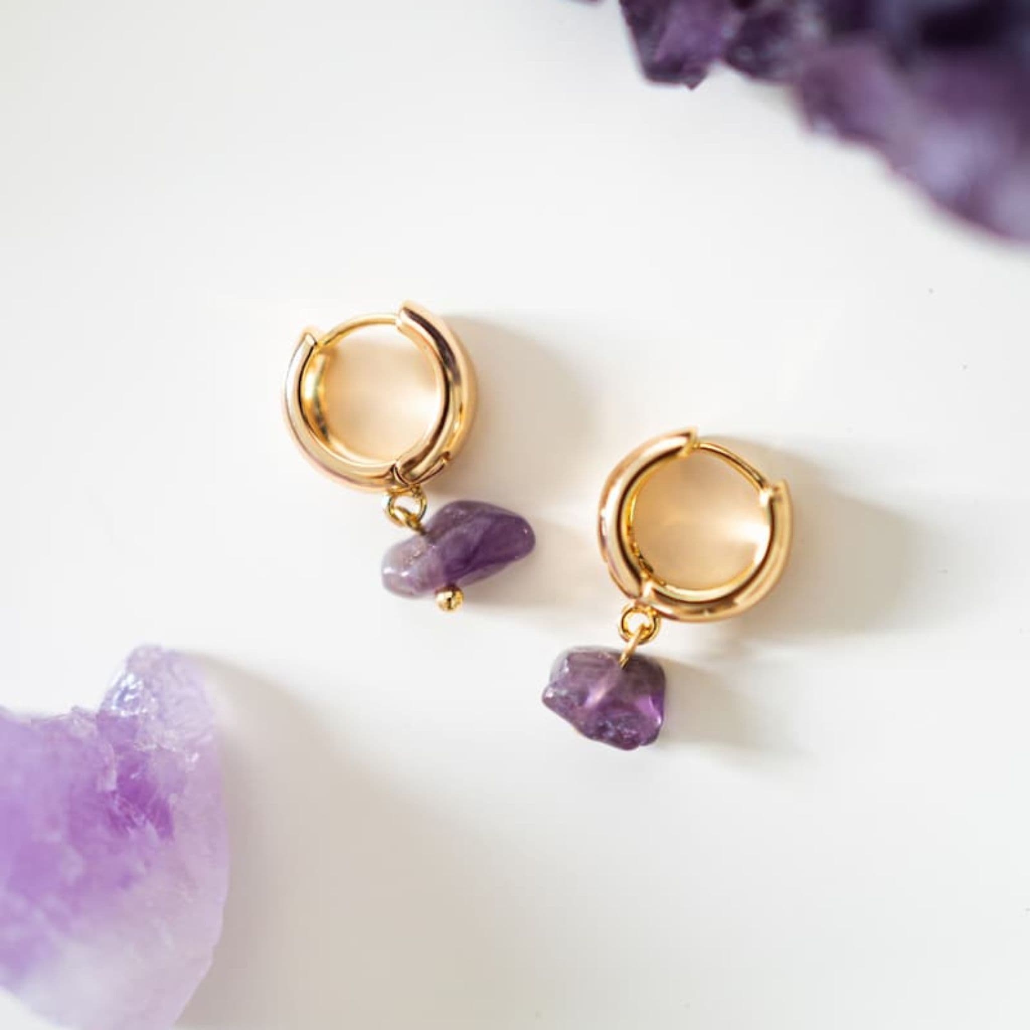Gold hoop earrings with purple gemstones on a white surface with purple stones.