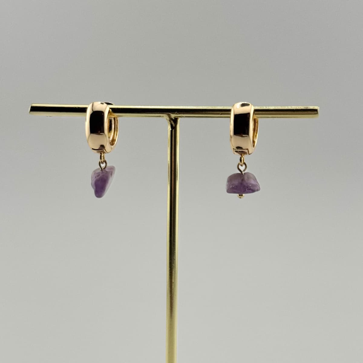 Gold hoop earrings with purple gemstones on a gold stand against a gray background