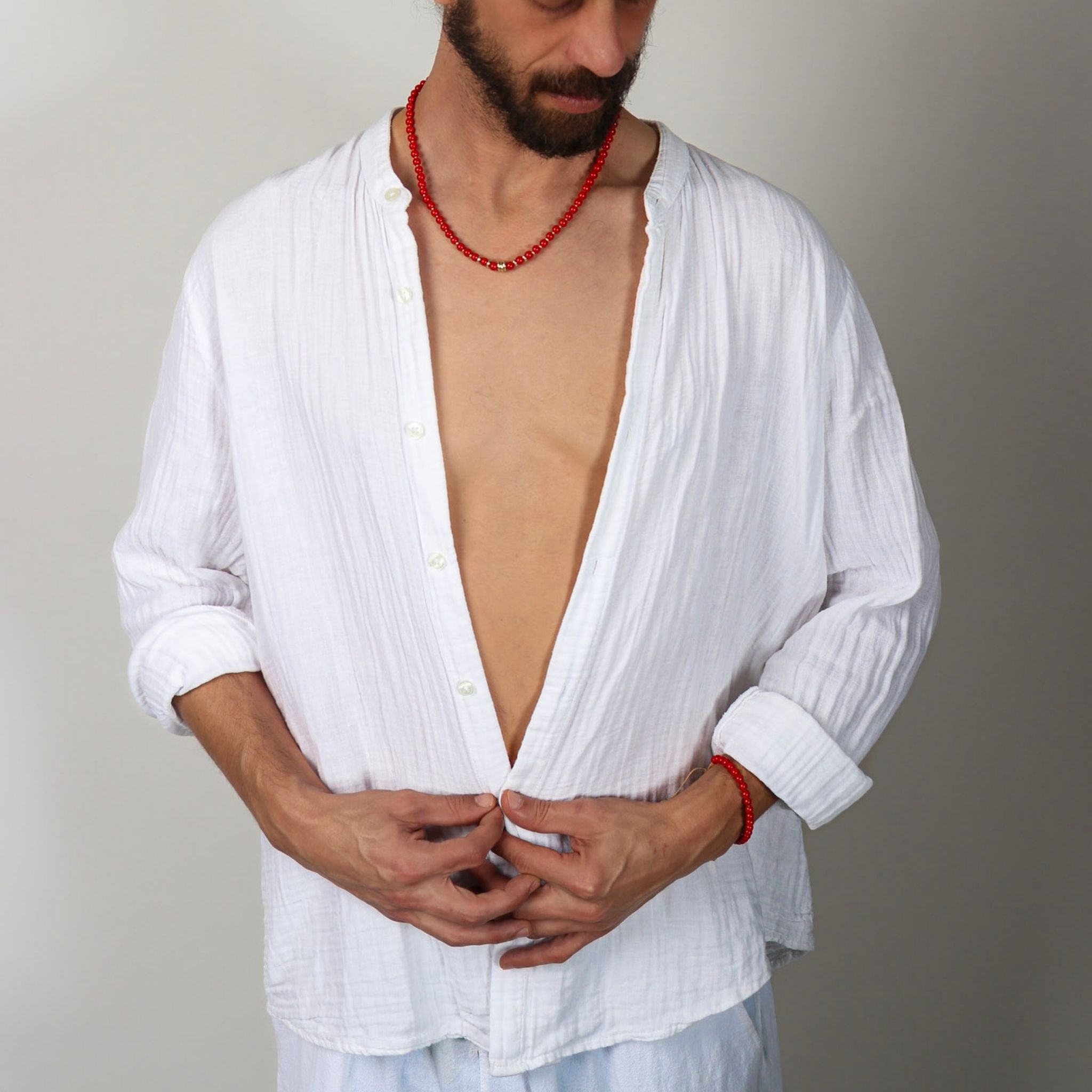 Man wearing a white shirt with open collar and red beads around his neck against a plain background