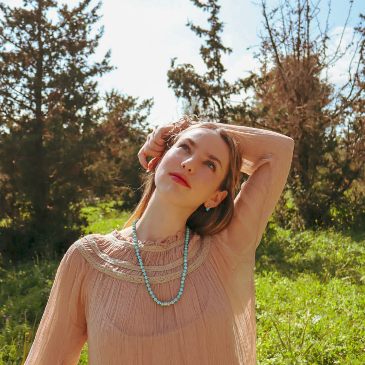 Woman in a pink dress with a blue necklace standing in a grassy field with trees in the background.