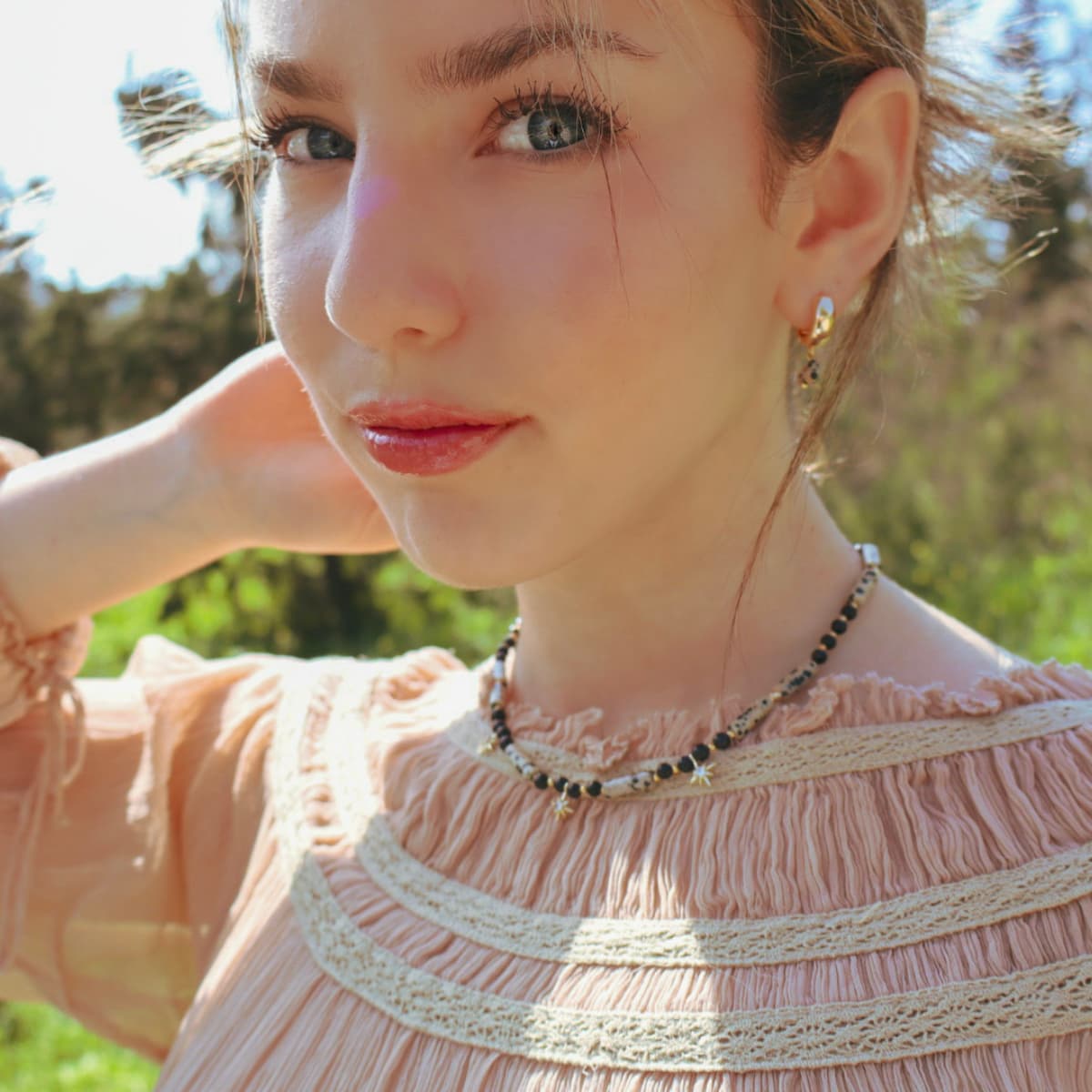 Woman wearing a pink top with lace details and layered necklaces outdoors.