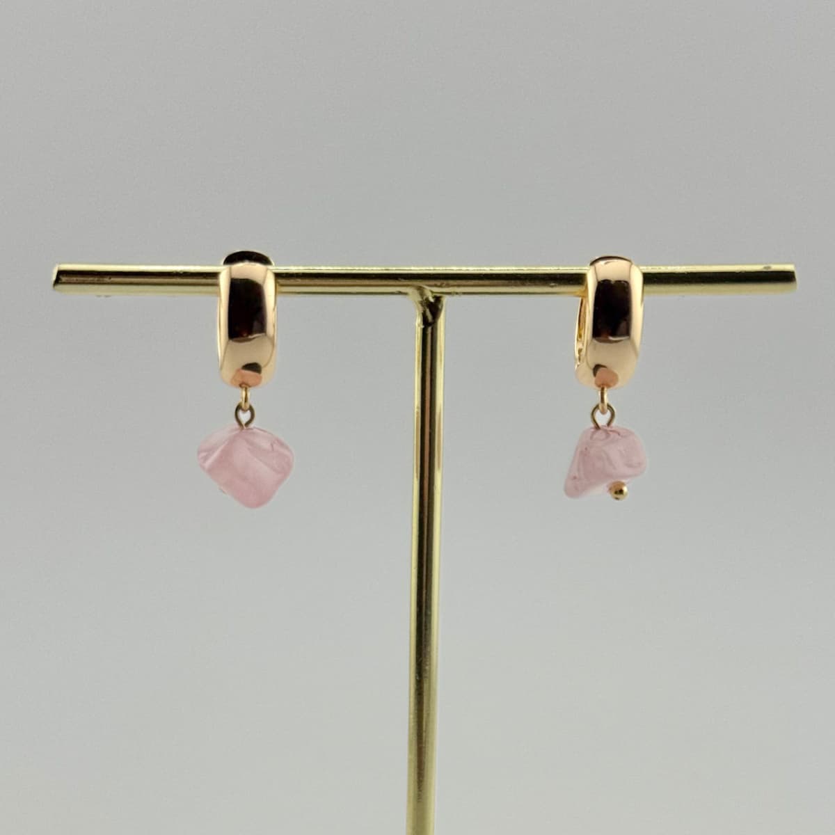 Gold hoop earrings with pink heart-shaped charms on a gold stand against a gray background